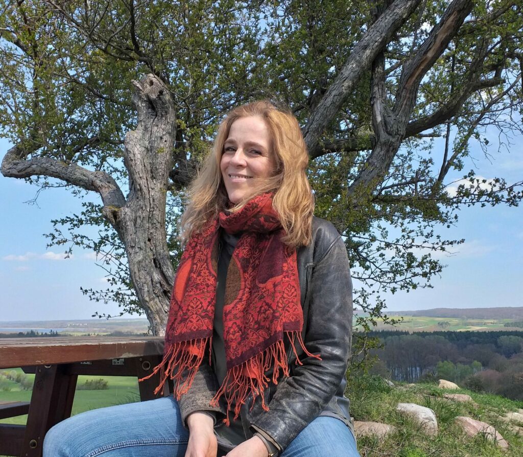 Fun Facts über Elena Deppe. Sie sitzt lachend auf einer Bank vor einem Baum.