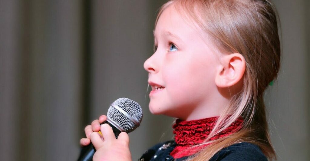 Ein Mädchen hält ein Mikrofon in den Händen. Sie blickt fragend nach oben und singt.
