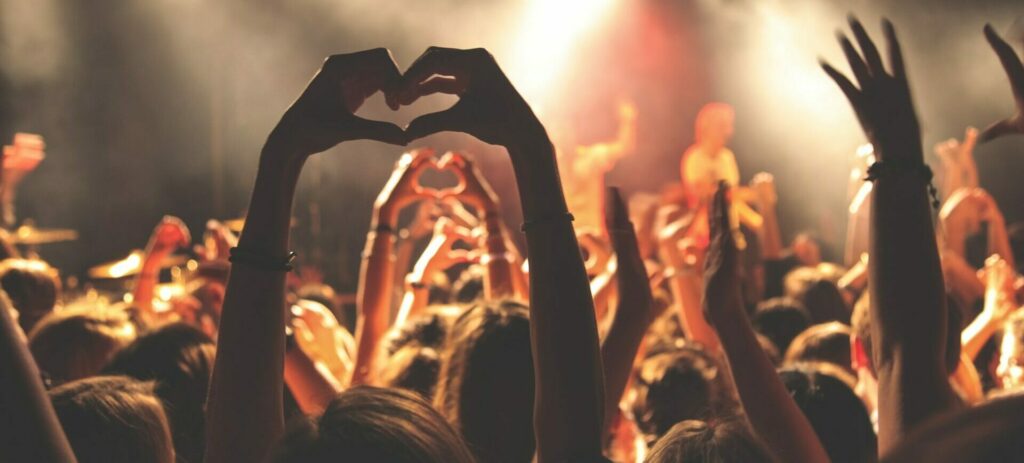 Viele Arme die sich hochstrecken. Hände formen Herzsymbole. Im Hintergrund eine Bühne mit Sänger und Band.
