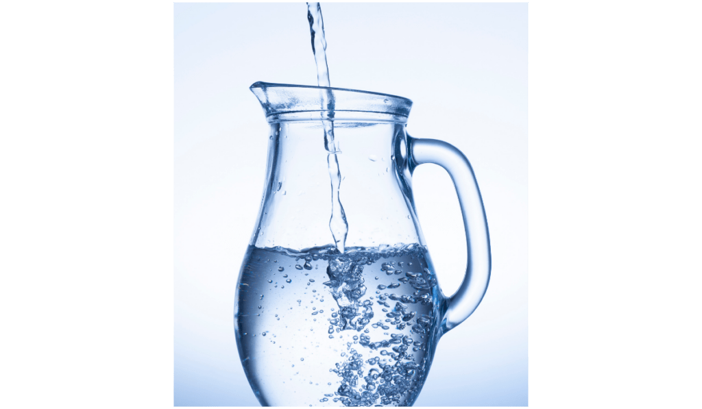 Ein Wasserkrug aus Glas wird mit Wasser befüllt. Er ist noch halbvoll. Der Wasserstrahl von oben verursacht Luftbläschen im Krug.