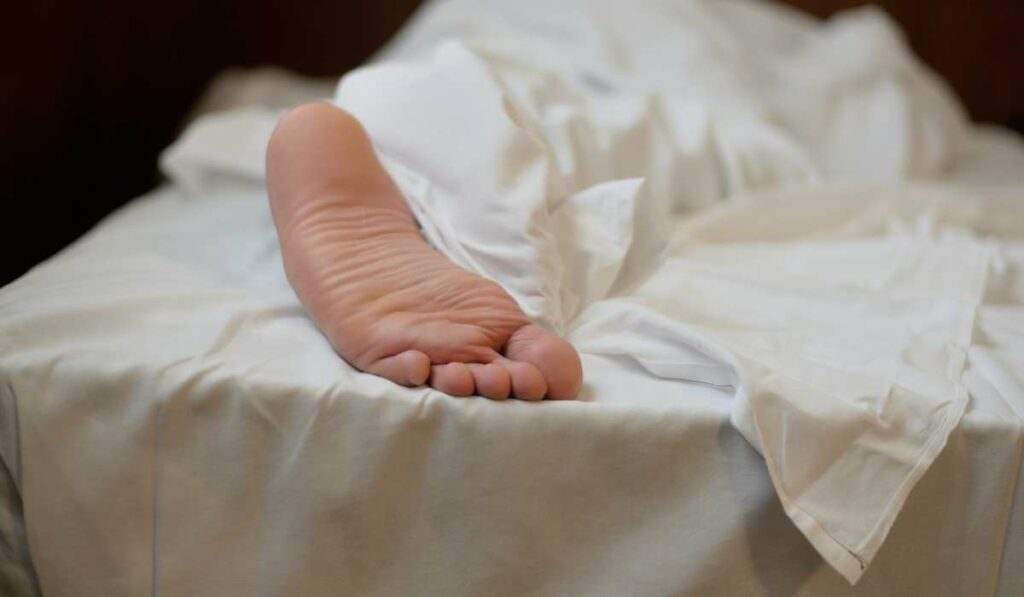 Das frühe Aufstehen bei der Morgenroutine fällt oft schwer. Ein nackter Fuß schaut aus unter der weißen Bettdecke heraus.